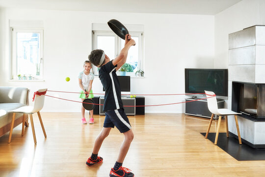 Siblings playing tennis with frying pans in living room at home during quarantine