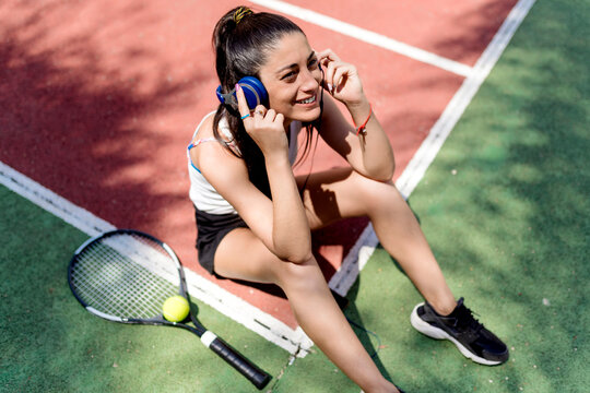 Smiling female tennis player listening music while sitting on floor in court