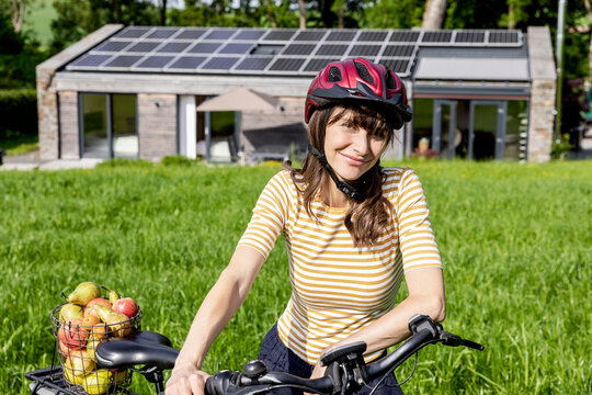 Portrait of smiling woman with bicycle and organic fruit on a meadow in front of a house