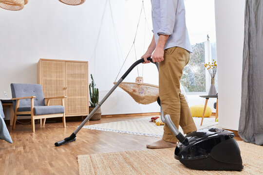 Baby boy on swing looking at father cleaning living room with vacuum cleaner