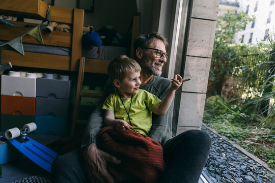 Father and little son sitting on the floor at home looking out of window