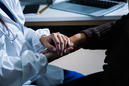 Senior patient lying in hospital bed receiving comforting touch from young female nurse, hands gently clasped together, medical care setting visible