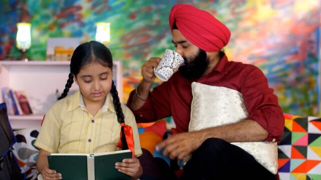 An Indian father helping his daughter with school homework - father-daughter bonding  book reading  me time. A Sikh Indian man helping his little kid with her studies while drinking a cup of hot te...