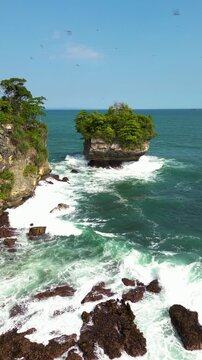 Vertical drone footage capturing powerful waves crashing against rocky shoreline and offshore islet at Karang Bokor Beach, Sawarna, Banten. Created a dynamic seascape scene.