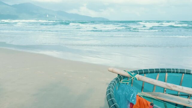 Coracle basket boat resting on my khe beach in da nang, vietnam, waves breaking against the sand and coastal mountains under a cloudy sky, representing traditional fishing and travel