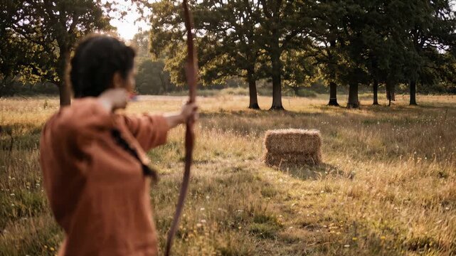 A person stands in a grassy field with trees in the background, preparing to shoot an arrow at a target. The focus is on the stance and technique of using a bow in natural light
