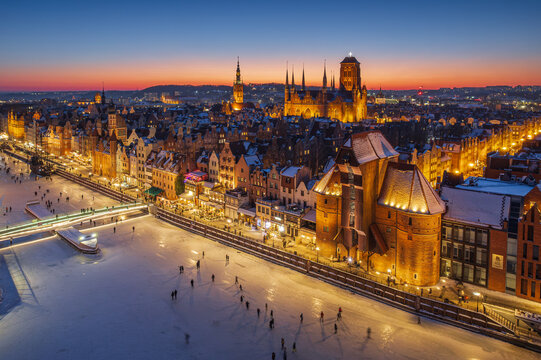 Aerial view of Gdansk Old Town at night in winter. Frozen Motlawa river, illuminated historic architecture, pedestrian bridge, and St. Mary's Basilica.