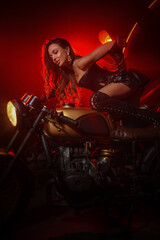 Fototapeta na wymiar Stylish beautiful young woman in black leather corset and boots posing on vintage motorcycle on old garage dark interior background.