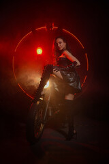 Naklejka na ściany i meble Stylish beautiful young woman in black leather corset and boots posing on vintage motorcycle on old garage dark interior background.