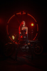 Fototapeta na wymiar Stylish beautiful young woman in black leather corset and boots posing on vintage motorcycle on old garage dark interior background.