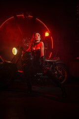 Fototapeta na wymiar Stylish beautiful young woman in black leather corset and boots posing on vintage motorcycle on old garage dark interior background.