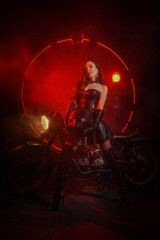 Naklejka na ściany i meble Stylish beautiful young woman in black leather corset and boots posing on vintage motorcycle on old garage dark interior background.