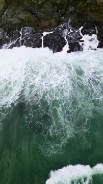 Vertical drone footage capturing a powerful ocean wave breaking over coastal rocks at Karang Bokor Beach, Sawarna, Indonesia. Rich turquoise water and white foam create a dramatic tropical seascape.