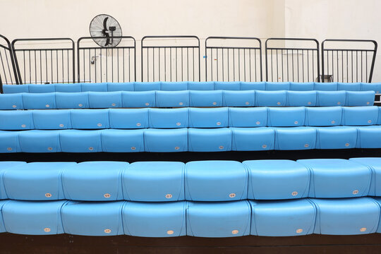 Blue stadium seats in empty indoor arena with black safety railings and wall fan, sport bleachers background for events and spectators.