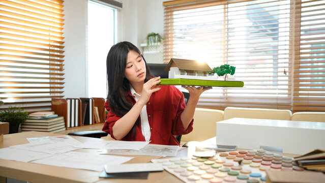 A female architect holding and working on an architecture house model with shop drawing paper in the office, Young woman architect building model house working in modern office