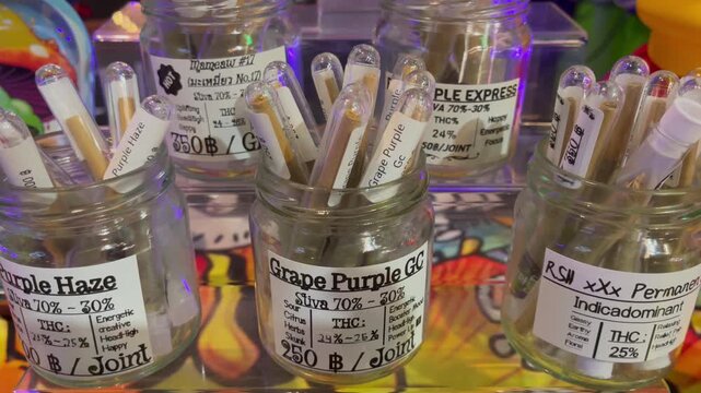 Close-up of labeled cannabis pre-rolled joints displayed in glass jars at a dispensary, showcasing different strains, THC levels, and sativa&ndash;indica profiles