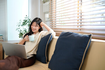Obraz premium A woman smiles while sitting and relaxing on a couch in a living room, holding a mug and using laptop