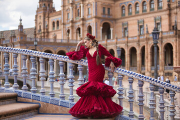Fototapeta premium Young, beautiful, blonde woman in red flamenco dress, flowers in her hair and gold jewelry, performing flamenco poses on a nice bridge in the square of Spain in Seville. Dance concept, art, typical.