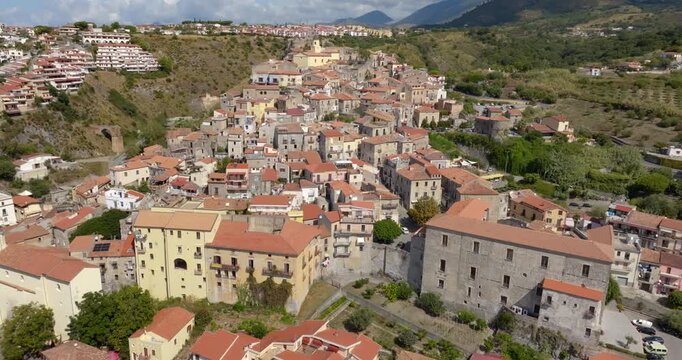 Aerial view of a historic Mediterranean village nestled on a lush hillside. There are many terracotta roofs, ancient stone architecture, and winding alleys. It is the town of Scalea, Calabria, Italy.