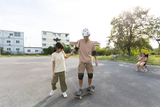 Teenage Asian Boy Practicing Skateboarding While Holding Younger Sister&rsquo;s Hand Outdoors