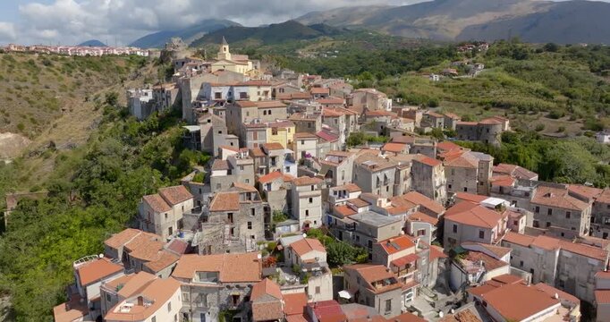 Aerial view of a Mediterranean hillside village, featuring a dense cluster of houses that climb toward a ridge, set against a backdrop of green hills and distant mountain peaks. It's Scalea, Italy.