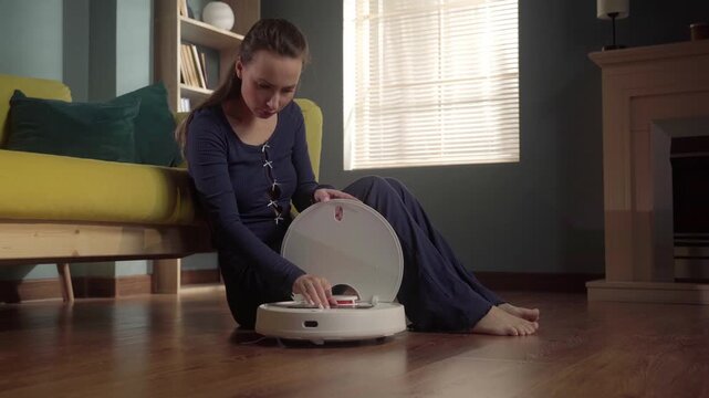 Woman sitting on a wooden floor in a cozy living room, opening the top cover of a robot vacuum cleaner, preparing for automated cleaning and smart home maintenance