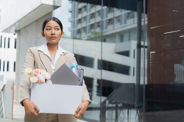 Stressed young woman in a suit carrying her personal items in a box while leaving an office building. Concept of unemployment, resignation, and career crisis.
