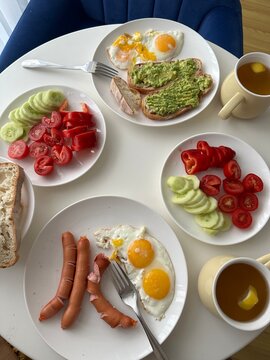 Breakfast spread on white table, fried eggs, sausages, avocado toast, fresh vegetables and lemon tea, top view flat lay, healthy morning meal for two.