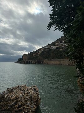 Seaside fortress and long stone wall below a hillside town on a calm bay under dramatic cloudy sky, historic coastal landmark and scenic travel destination with copy space.