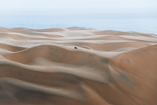 View of a lone vehicle braving the undulating dunes where golden sand meets the distant blue of the ocean horizon, Walvis Bay, Erongo Region, Namibia.
