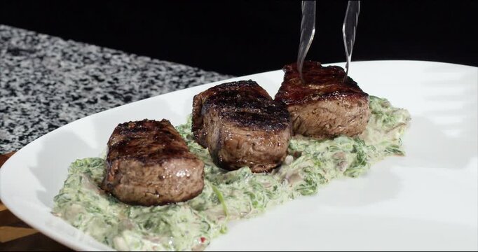 Placing beef steaks onto pillow of garlic rosemary sauce closeup. Elegant serving of delicacy veal dish with spicy seasoning in restaurant kitchen. Fine dining