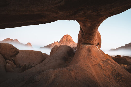 View of massive granite boulders, sculpted by time and elements, frame a misty valley and distant peaks under a soft, ethereal light, Spitzkoppe, Erongo Region, Namibia.