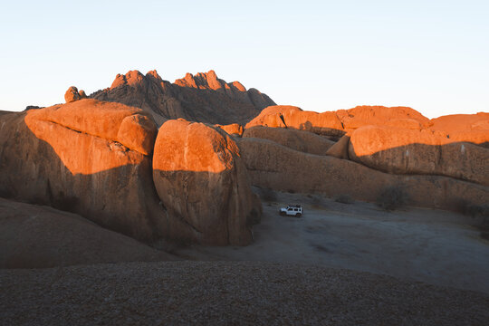 View of warm sunlight bathing the rugged, rocky landscape where an isolated vehicle sits nestled among the massive boulders, creating a striking contrast of human presence in Namibia.