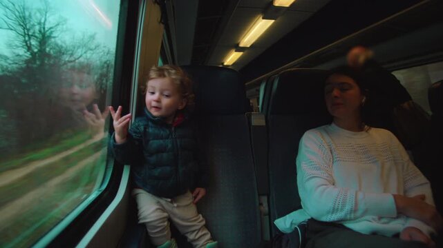 Toddler standing on train seat pointing at passing countryside through window while parent rests nearby evoking curiosity shared travel bonding attention early memory and quiet discovery