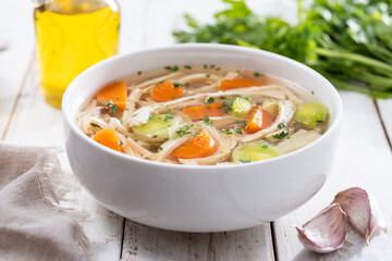 Homemade chicken soup in white bowl with carrots and celery on white wooden table