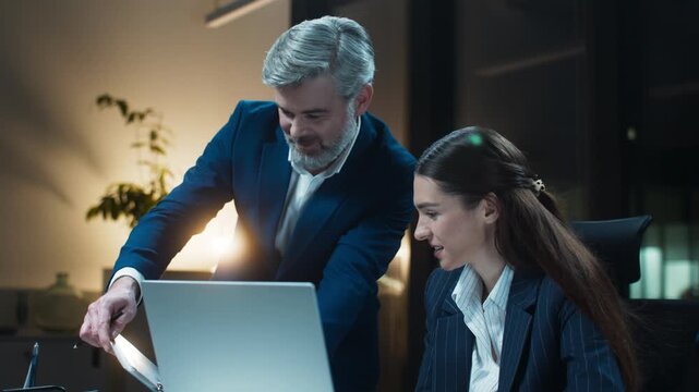 Business discussion at desk during late hours. Caucasian man standing and pointing. Female manager seated and listening. Explaining chart improvements. Sharing positive results and progress.