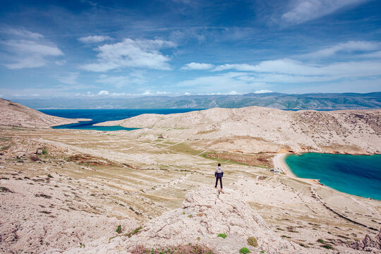 View of a solitary figure stands atop a barren, rocky landscape overlooking the turquoise Adriatic Sea under a vast sky, Baska, Primorje-Gorski Kotar County, Croatia.