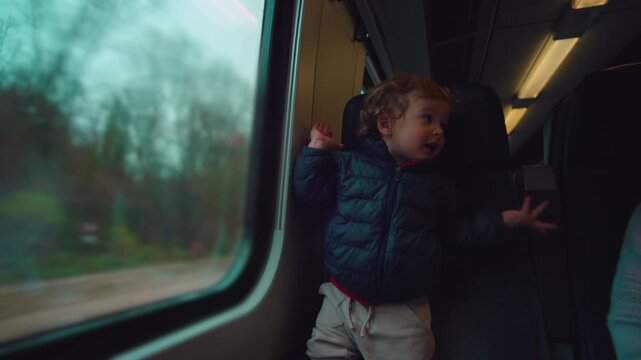 Child standing on train seat watching outside world pass by window with absorbed expression as rural landscape slides past suggesting curiosity, imagination, quiet joy, travel intimacy