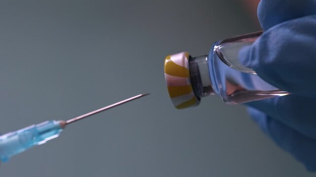 Extreme close-up of a needle piercing a vial stopper while a gloved hand pulls the syringe plunger to draw liquid medication. Medical preparation and vaccination concept.