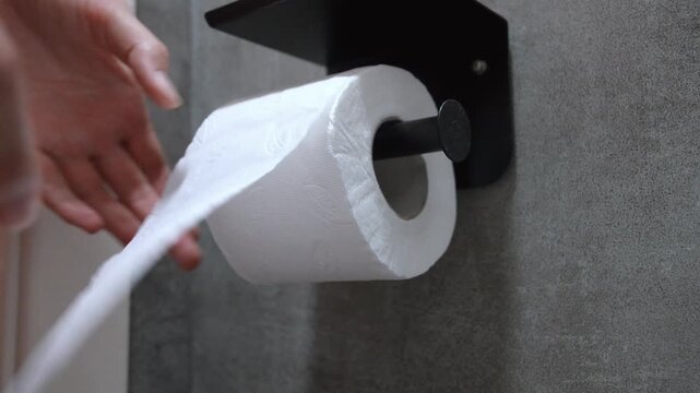 Close-up view of a person's hand pulling and tearing a sheet from a white toilet paper roll, which is mounted on a modern black holder against a textured gray bathroom wall