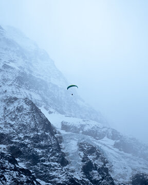 View of a lone paraglider soars against a backdrop of stark, snow-dusted mountain peaks cloaked in a misty veil, Airolo, Ticino, Switzerland.