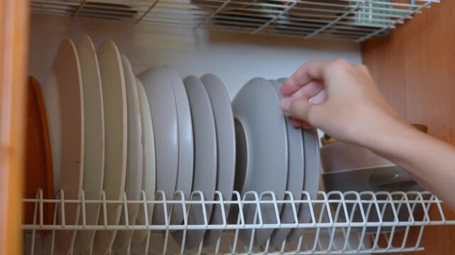 Female hand taking a clean grey plate from a white metal dish drying rack inside a wooden kitchen cabinet, preparing for a meal or setting the table during daily household chores at home