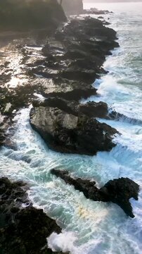 Aerial drone footage capturing warm sunlight reflecting on wet rock surfaces and flowing ocean water at Sawarna Beach, Indonesia. Natural coastal contrast highlighted in immersive Vertical frame.