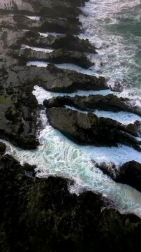 Vertical drone shot showing ocean waves streaming between long rocky ridges at Karang Taraje Beach, Indonesia. Detailed textures of dark rocks and turquoise water create a powerful natural seascape.
