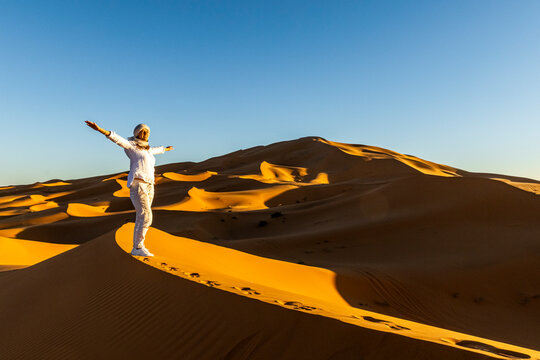 A woman in white stands with arms outstretched on a sand dune, under a clear blue sky. Mezouga,Sahara desert,Morocco