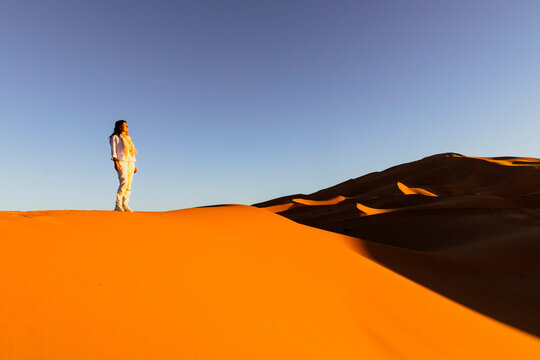 A woman in white stands atop a vibrant orange sand dune, gazing towards the horizon under a clear blue sky. Mezouga,Sahara desert,Morocco