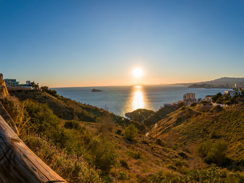 View of the radiant sun setting over the tranquil sea, casting a golden glow on the hills and distant cityscape, Benidorm, Valencian Community, Spain.