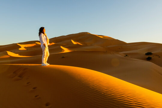 A woman in white stands atop a sand dune, gazing at the vast desert landscape under a clear sky. Mezouga,Sahara desert,Morocco