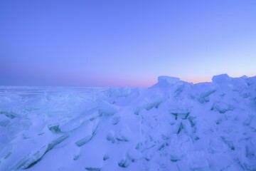 A frozen shoreline covered with jagged ice formations and snow under a soft twilight sky. The horizon glows with gentle pink and orange tones fading into cool blue, suggesting sunrise or sunset. © Gala_Rocci