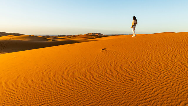A woman stands atop a large sand dune, gazing at the vast desert landscape under a clear sky. Mezouga,Sahara desert,Morocco